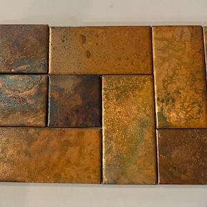 May include: A collection of copper tiles with a distressed finish. The tiles are in various sizes and shapes, including rectangular and square. The colours range from copper and bronze to teal and brown.