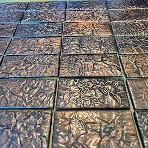 May include: A close-up of a sheet of metallic tiles. The tiles are a dark brown color with a textured surface that resembles crumpled foil. The tiles are arranged in a grid pattern.