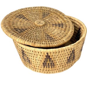 Op de afbeelding: Een ronde rieten mand met een deksel. De mand heeft een natuurlijke kleur en een geometrisch patroon in donkerbruin. Het deksel is gedeeltelijk open en toont de binnenkant. Deze decoratieve mand is een opbergcontainer.