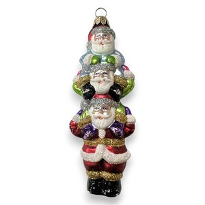 May include: A Christmas tree ornament depicting three Santa figures, one on top of the other. The ornament has a gold cap and is painted with red, blue, green, and gold glitter accents. The Santas have white beards and black boots.