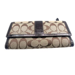 May include: A brown and tan Coach wallet with a repeating logo pattern. The wallet has a dark brown leather strap and trim. The wallet is closed and has a snap closure.