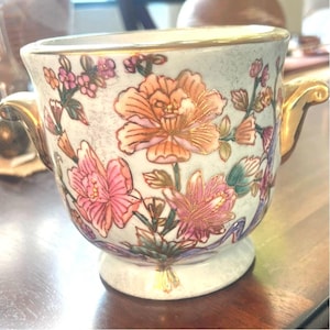 Peut inclure: Une tasse décorative en céramique avec un motif floral. La tasse a une base blanche avec des accents dorés sur les poignées et le bord. Le motif floral comprend des fleurs roses et oranges avec des feuilles vertes.