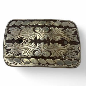 Puede incluir: Una hebilla de cinturón plateada con fondo marrón y un diseño floral decorativo.