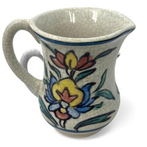 Puede incluir: Una jarra de cerámica pequeña, de color blanquecino, con asa y pico vertedor. La jarra está decorada con un diseño floral en amarillo, azul, rojo y verde. El borde está adornado con una banda azul. La jarra tiene un esmalte craquelado.