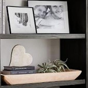 Puede incluir: Una estantería de madera exhibe fotos en blanco y negro enmarcadas, libros y objetos decorativos. Un corazón de madera de color claro se encuentra encima de dos libros oscuros. Un cuenco de madera contiene pequeñas plantas verdes.