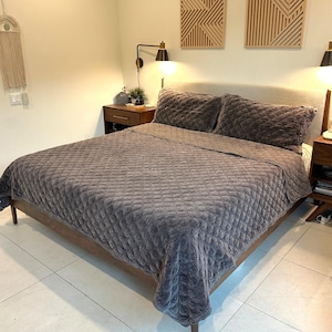May include: A bed with a grey, textured quilt and matching pillows. The bed frame is made of wood. The room has wooden wall art and sconce lighting.