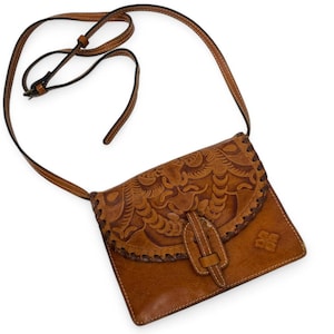 May include: A brown leather crossbody bag with a floral embossed design on the flap. The bag features a long, adjustable strap and a buckle closure. The bag has a stitched border and a small logo on the lower right corner.