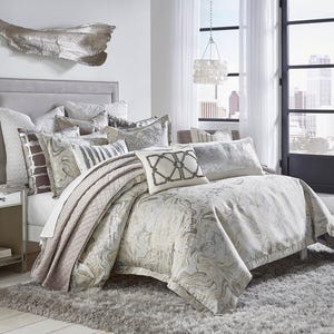 May include: A luxurious bedroom setting featuring a bed with a gray upholstered headboard and a plush, patterned comforter. Decorative pillows in various textures and patterns add to the elegant aesthetic. A mirrored nightstand and a fluffy gray rug complete the scene.