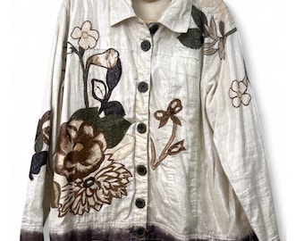 May include: Cream-coloured button-up jacket with floral appliqué. The jacket features a shimmering fabric with brown, green, and cream-coloured floral designs. The jacket has a collar and dark brown buttons. The bottom hem has a darker brown trim.