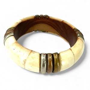 May include: Cream-colored bangle bracelet with a dark brown interior and metal accents. The bracelet features rectangular segments and bands of silver, bronze, and gold-toned metal. A unique piece of jewelry.