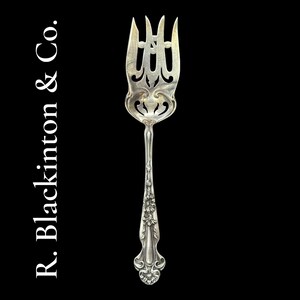 Può includere: Una forchetta da portata d'argento con un design ornato. La forchetta ha quattro rebbi con intagli decorativi e un motivo floreale sul manico. Il testo "R. Blackinton & Co." è stampato verticalmente sul lato sinistro dell'immagine.