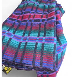 Puede incluir: Una manta doblada con un estampado de cuadros en tonos turquesa, morado, rojo y negro. La manta tiene una etiqueta con texto dorado. La manta está hecha de un material suave.