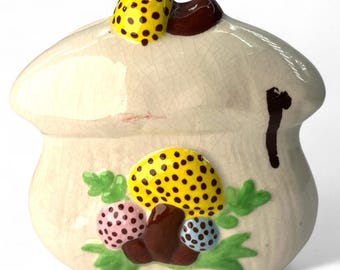 May include: A ceramic mushroom-shaped container with a cream-colored base and a lid. The container is decorated with yellow mushrooms with black spots, pink and blue mushrooms, and green leaves. The lid has a yellow mushroom with black spots and a brown accent.
