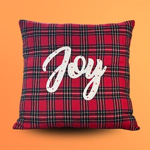 以下が含まれることがあります： 赤と黒のチェック柄の正方形の装飾枕。「Joy」の文字が白い筆記体で前面に書かれており、小さな赤い装飾が施されています。お祝いのホリデーデコレーションです。