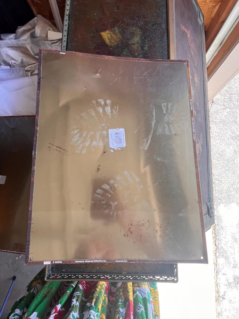 May include: A rectangular metal sign with a gold background and a brown border. The sign has visible wear and tear, including scuffs and marks. The bottom left corner has the Marvel logo and the words "Licensed to Desperate Enterprises, Inc."