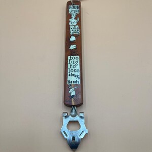 May include: A vintage bottle opener with a wooden handle and a metal opener. The handle is brown with white text and illustrations, including "FOUR tools in one" and "too big to lose always Handy". A string is attached at the top.