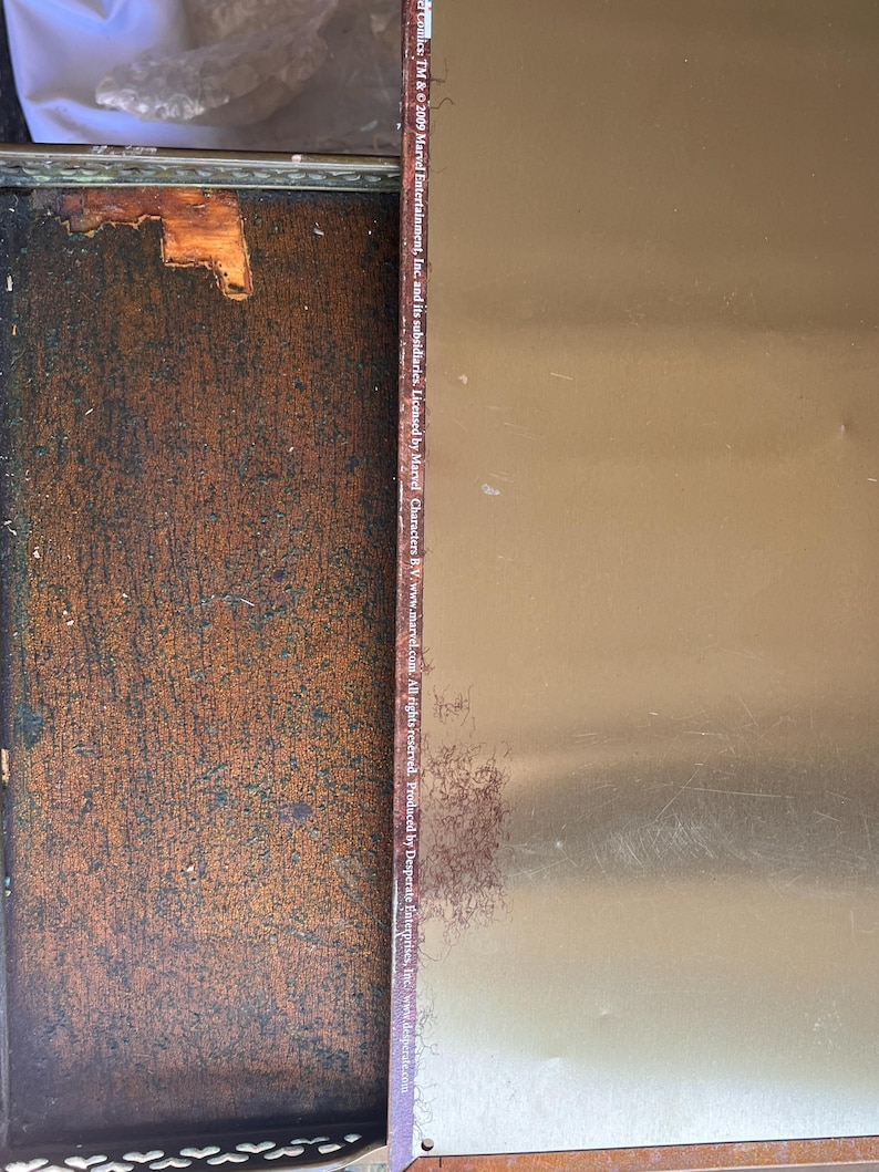 May include: A metal sign with a gold-colored surface and text from Marvel Comics. The sign has some rust and wear. The text includes copyright information and the Marvel website address.