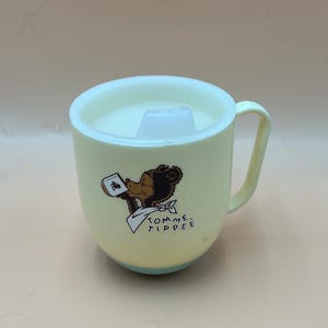 May include: A yellow Tommee Tippee sippy cup with a white lid and handle. The cup features a cartoon bear illustration holding a card and the words "Tommee Tippee". The base of the cup is light blue.