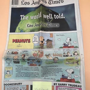 May include: A folded Los Angeles Times newspaper featuring the Peanuts comic strip. The headline reads "The world well told." The comic strip includes characters like Charlie Brown and Snoopy. A yellow sticky note says "Last Peanuts."