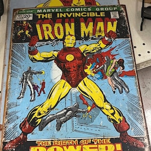 May include: Vintage metal sign featuring the Iron Man comic book cover. The artwork shows Iron Man in a red and yellow suit, with the text "IRON MAN" in large letters. The sign has a distressed, aged appearance.