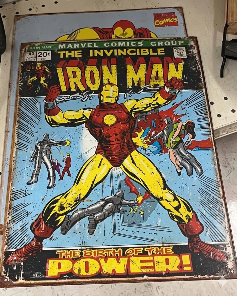 May include: Vintage metal sign featuring the Iron Man comic book cover. The artwork shows Iron Man in a red and yellow suit, with the text "IRON MAN" in large letters. The sign has a distressed, aged appearance.