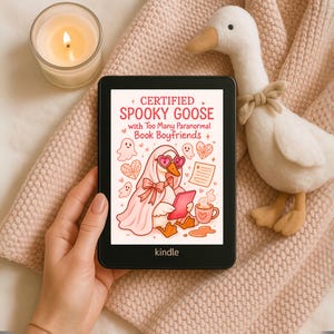 May include: A Kindle e-reader displays a cartoon illustration of a goose in a ghost costume, with the text "Certified Spooky Goose with Too Many Paranormal Book Boyfriends." A plush goose and a lit candle are nearby.