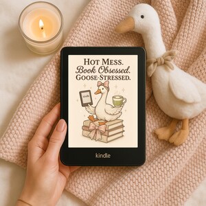 May include: A black Kindle e-reader displaying the text "Hot Mess. Book Obsessed. Goose-Stressed." The screen shows a cartoon goose holding a Kindle and a cup of tea, sitting on books. A plush goose and a lit candle are nearby.