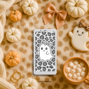 May include: A white smartphone with a gray leopard print background and a cartoon ghost with a bow. The image includes pumpkins, a bow, a ghost-shaped cookie, a mug of hot chocolate, and gold rings, all on a tan spotted blanket.