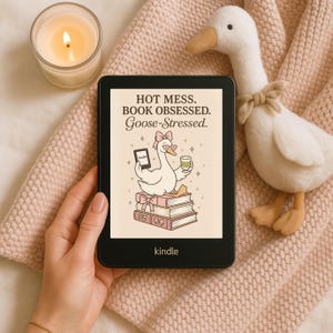 May include: A black Kindle e-reader displays a cartoon goose illustration with the text "HOT MESS. BOOK OBSESSED. Goose-Stressed." A plush goose toy and a lit candle are in the background. The e-reader is held by a person.