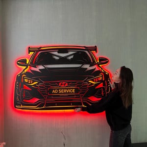 May include: Black car-shaped wall art with red LED lighting. The artwork features the front of a car with the text "AD SERVICE" on the front. The car has a spoiler and detailed headlights. The art is mounted on a gray wall.
