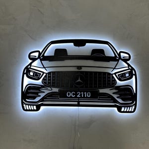 May include: Black and white car silhouette with illuminated edges. The car features a prominent front grille with the Mercedes-Benz logo and the license plate reads "OC 2110". The design is mounted on a gray wall.