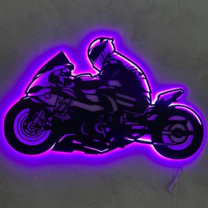 Puede incluir: Silueta negra de una motocicleta con un piloto, iluminada por una vibrante luz LED púrpura. La motocicleta tiene el número 77. La obra de arte está montada en una pared gris, creando un contraste llamativo.