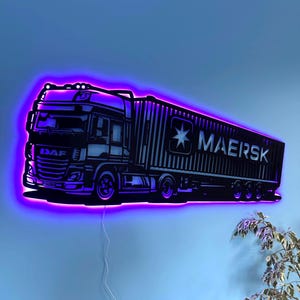 Puede incluir: Una obra de arte mural iluminada en negro y morado que representa un camión y un remolque. El remolque presenta la palabra "MAERSK" y un logotipo de estrella. El camión tiene el logotipo "DAF". El arte está montado en una pared azul.