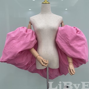 May include: A pink, high-low top with large, puffed sleeves is displayed on a mannequin. The sleeves are voluminous and extend outward, creating a dramatic silhouette. The mannequin has a gold-colored decorative top.