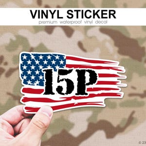 May include: A vinyl sticker featuring a distressed American flag design with the text "15P" in bold black letters. The sticker has a white border and is held against a camouflage background. The text "VINYL STICKER" is at the top.