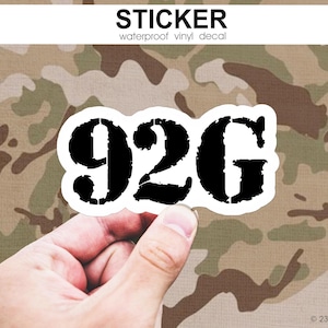 May include: A white sticker with the black text "92G" is held in front of a camouflage background. The sticker is a waterproof vinyl decal. The word "STICKER" is printed above the numbers.