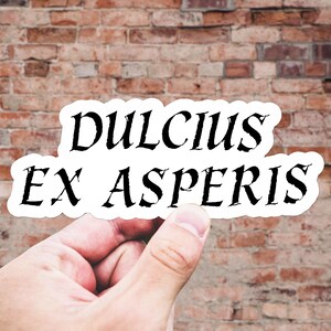以下が含まれることがあります： レンガの壁を背景に、手で持たれた、黒い文字で「DULCIUS EX ASPERIS」と書かれた白いステッカー。ステッカーは丸みを帯びた形で、テキストは装飾的なフォントです。