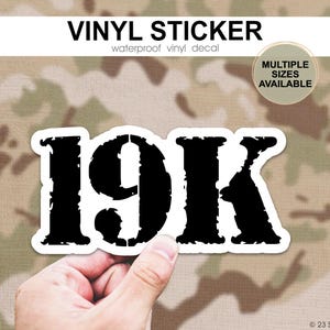 May include: A black and white vinyl sticker with the text "19K" in a distressed font. The sticker is waterproof and available in multiple sizes. The background is a camouflage pattern.