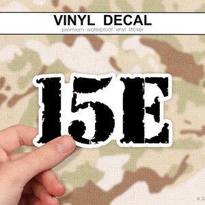 Könnte beinhalten: Ein schwarz-weißer Vinyl-Aufkleber mit dem Text "15E" in einer verwitterten Schriftart. Der Aufkleber wird von einer Hand vor einem Tarnmuster-Hintergrund gehalten. Oben stehen die Worte "VINYL DECAL".