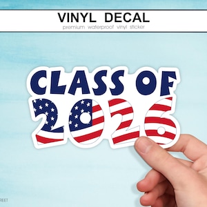 May include: A white vinyl sticker with the text "CLASS OF 2026" in navy blue and red, white, and blue American flag design. The sticker is held by a hand against a light blue background. The top of the sticker reads "VINYL DECAL".