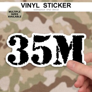 May include: A black and white vinyl sticker featuring the text "35M" in a distressed font. The sticker is set against a camouflage pattern background. The top of the image reads "VINYL STICKER" and "waterproof vinyl decal".