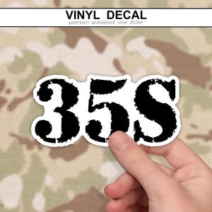 Peut inclure: Un autocollant en vinyle présentant le texte noir 35S dans une police de style vieilli et usé. L'autocollant a une bordure blanche et est présenté sur un fond à motif camouflage. Le texte VINYL DECAL et premium waterproof vinyl sticker figurent en haut.