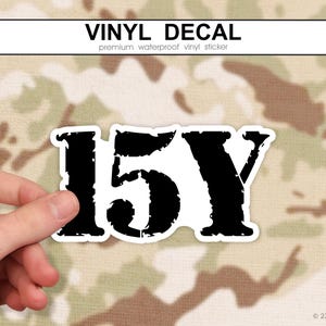 May include: A black and white vinyl decal with the text "15Y" in a bold, distressed font. The decal is held against a camouflage background. The words "VINYL DECAL" are printed at the top.