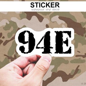 Könnte beinhalten: Ein schwarz-weißer Aufkleber mit dem Text "94E" in einer verwitterten Schriftart. Der Aufkleber wird von einer Hand vor einem Tarnmuster-Hintergrund gehalten. Das Wort "STICKER" ist über dem Aufkleber aufgedruckt.