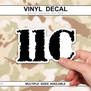 May include: Black and white vinyl decal with the letters IIC, featuring a distressed, textured design. The decal is held by a hand against a camouflage background. The text "VINYL DECAL" and "MULTIPLE SIZES AVAILABLE" are also visible.
