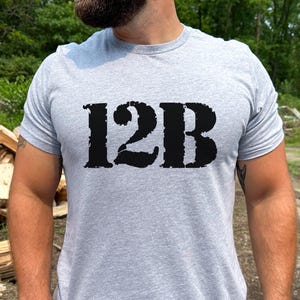 Puede incluir: Camiseta gris claro con el texto "12B" en letras negras grandes y desgastadas en el pecho. La camiseta tiene mangas cortas y cuello redondo. El fondo incluye una pila de leña y follaje verde.