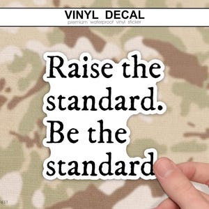 May include: A white vinyl decal sticker featuring the black text "Raise the standard. Be the standard." The sticker has a die-cut shape that follows the contour of the lettering, which is presented in a bold, serif font style for clear readability.