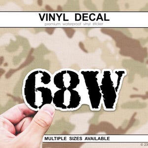 May include: A black and white vinyl decal with the text "68W" in a bold, distressed font. The decal is held against a camouflage background. The top of the image reads "VINYL DECAL".