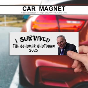 May include: A rectangular car magnet with the text "I SURVIVED THE SCHUMER SHUTDOWN 2025" and an image of a man. The magnet is held in front of a red car. The top of the magnet reads "CAR MAGNET".