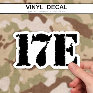 May include: A black and white vinyl decal with the text "17E" in a distressed font. The decal is held against a camouflage background. The top of the decal reads "VINYL DECAL".
